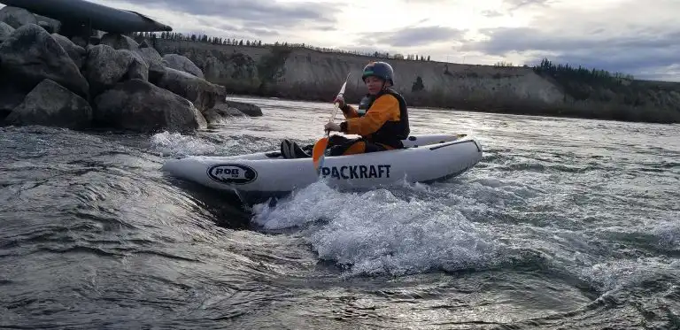 Paddler in a helmet and life jacket rides a white packraft through a wave below rocky banks