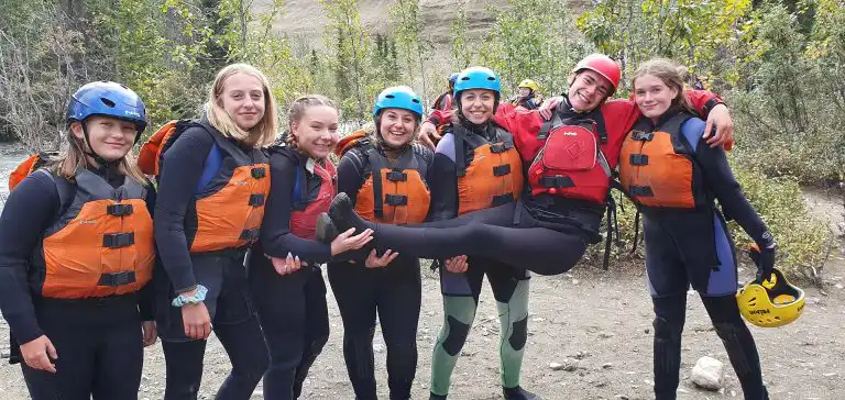 Group of smiling teens in wetsuits, helmets and life jackets poses on a riverbank