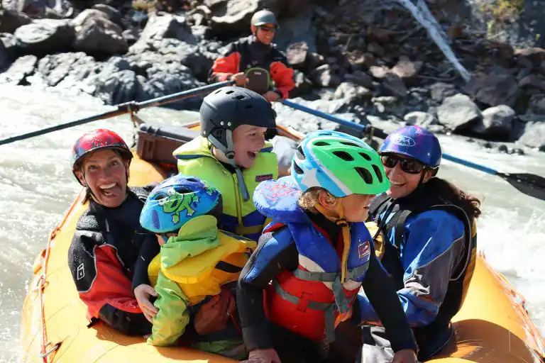 Smiling kids in helmets and life jackets ride a yellow raft with adults at Whitehorse kids camp
