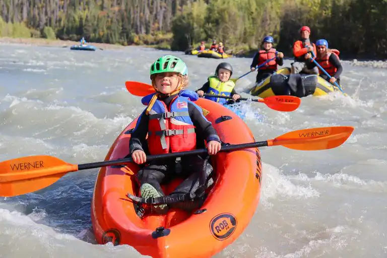 Young child paddling an orange inflatable kayak with rafts behind on a wide river